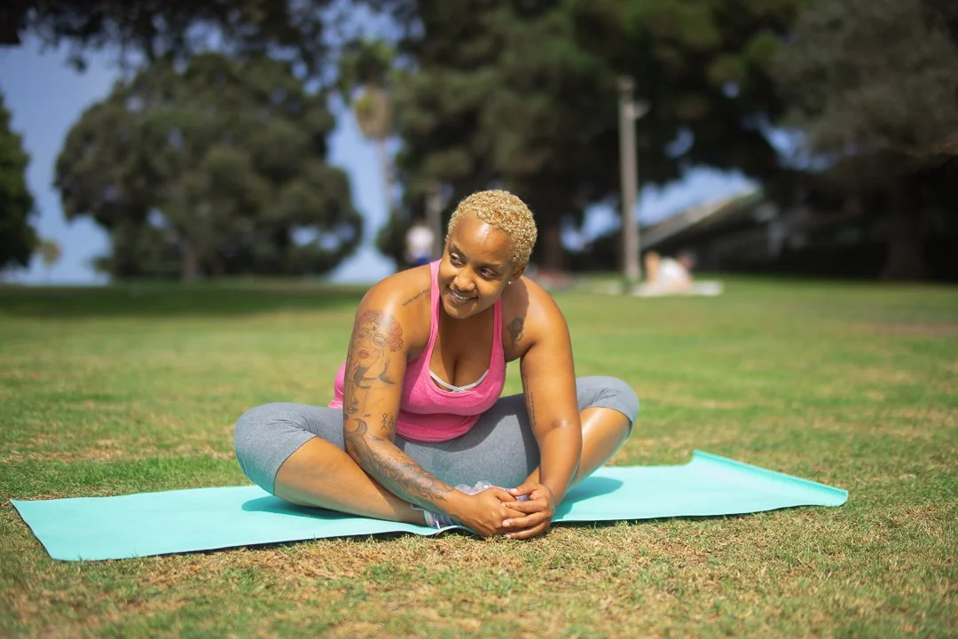 A woman practicing yoga outdoors on a mat in a park with trees and blue sky.