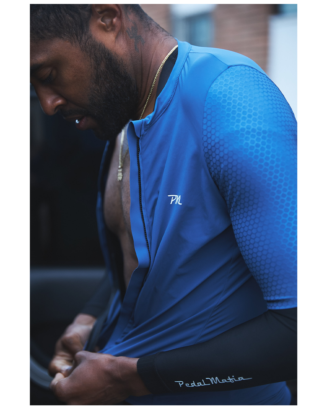 A man in a blue sports jacket with the initials 'PML' on the chest, adjusting his clothing, with a black undershirt and a gold chain around his neck.