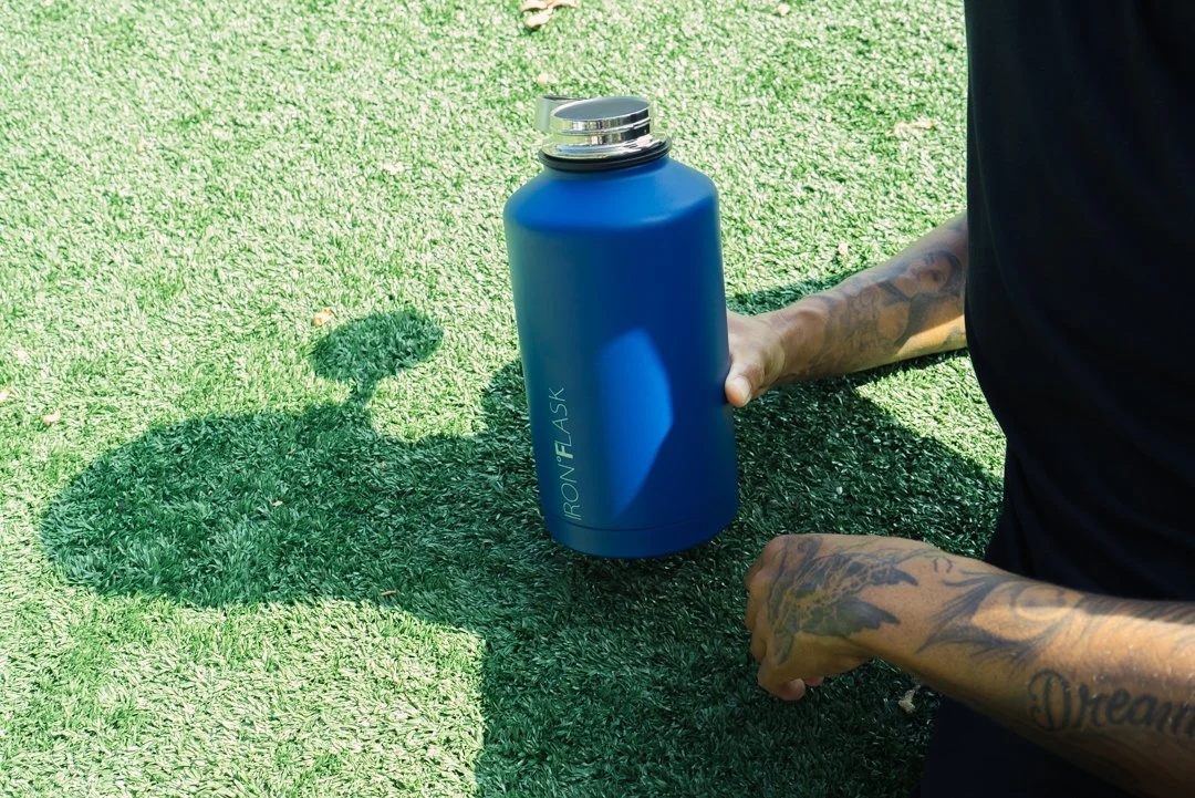 A person with tattoos on their arm holding a blue ION8 water bottle on grass, with their shadow cast on the ground.