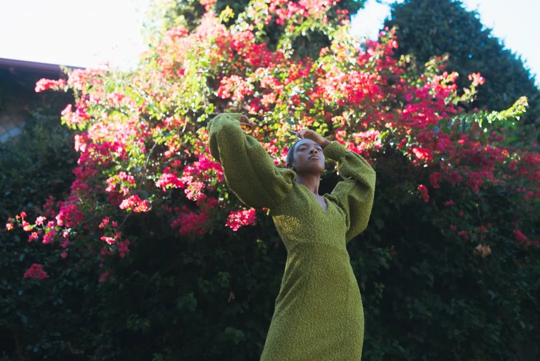 A woman in a green dress posing outdoors in front of blooming pink and red flowers with green foliage.