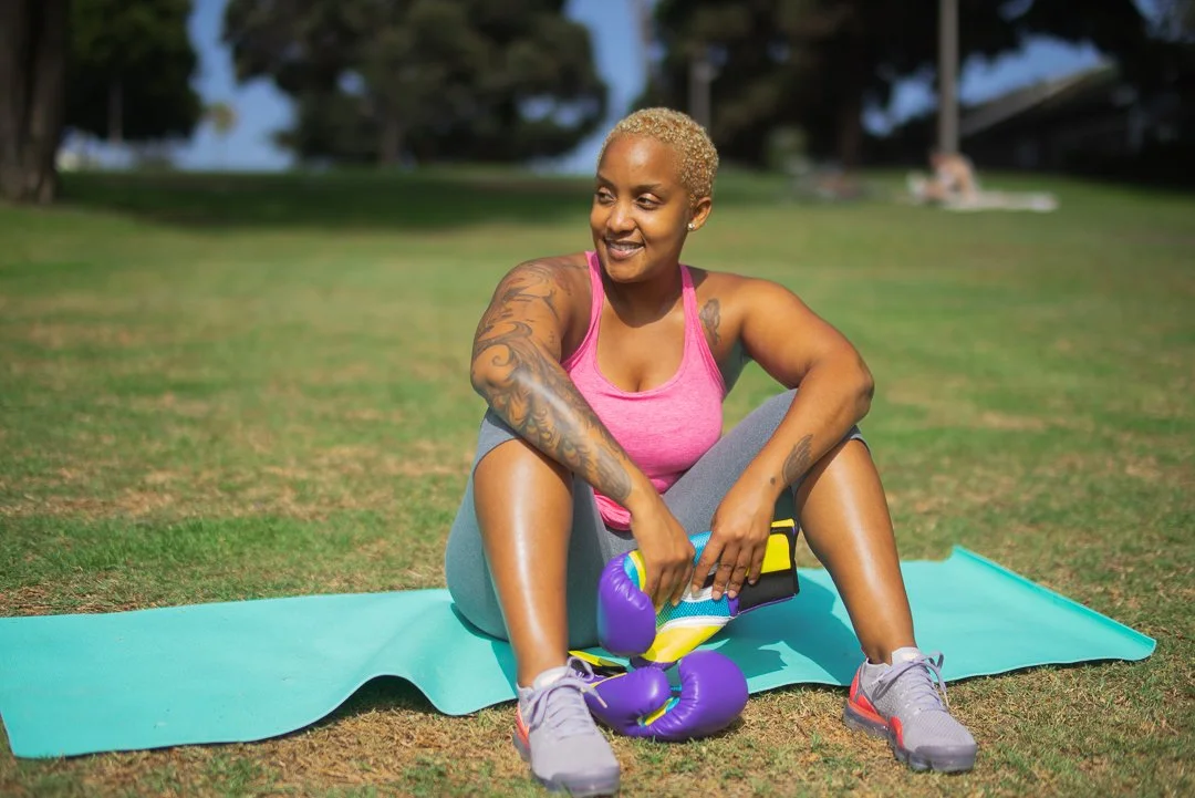 A woman with short blonde hair and tattoos sitting on a workout mat outdoors, holding purple boxing gloves, smiling, wearing a pink tank top and gray workout pants.