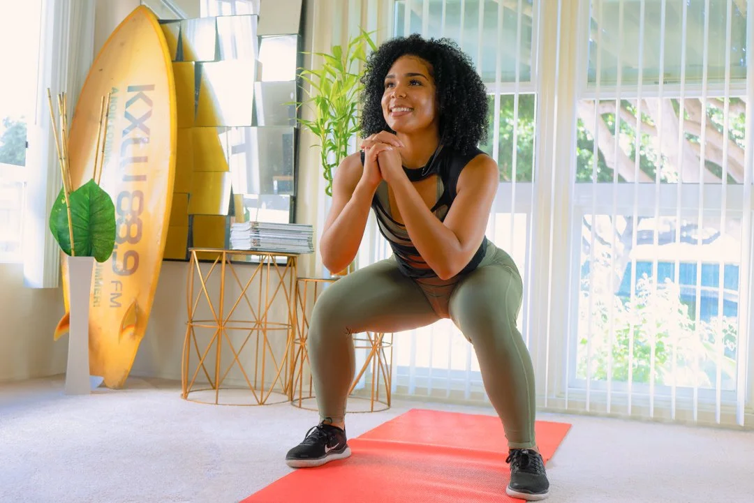 Woman performing squat exercise on a yoga mat in a bright room with surfboard, plants, and window with vertical blinds.