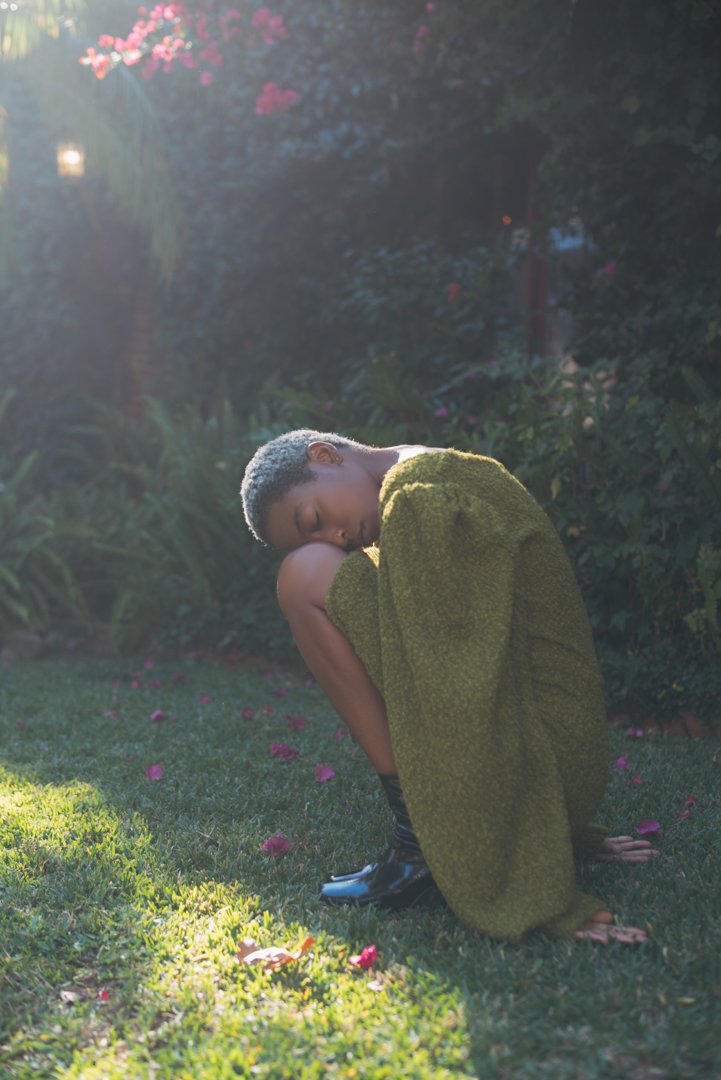 A woman with short, curly, gray hair crouches on a lawn, resting her head on her knee. She wears a green, textured coat, black socks, and shoes. The scene is lit by soft, natural sunlight, with flowering bushes and plants in the background.