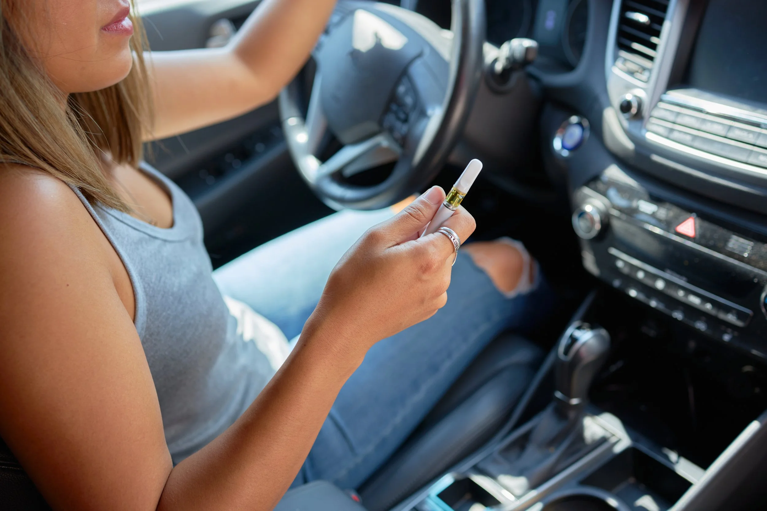 A woman sitting in the driver's seat of a car, holding a vape pen in her hand, with her left hand on the steering wheel.