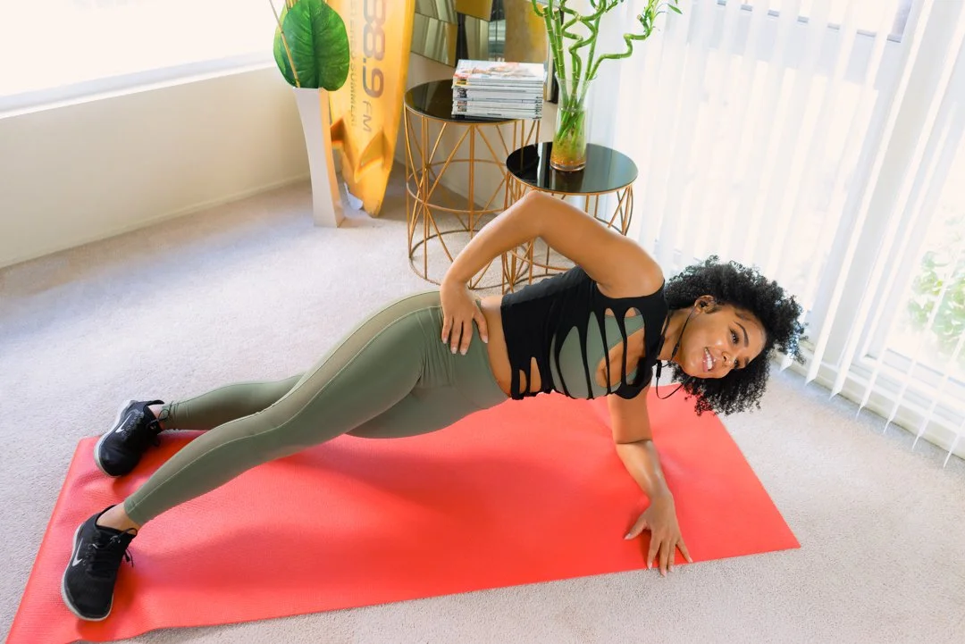 A woman exercising on a red yoga mat indoors, wearing black workout top and green leggings.