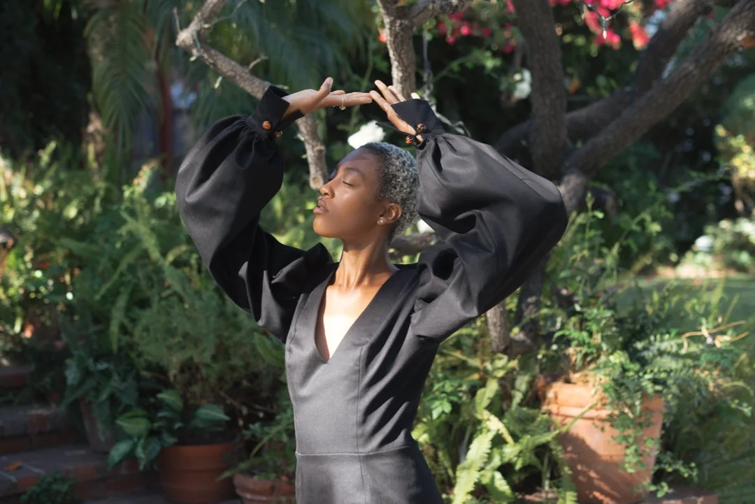 Woman with short gray hair wearing a black dress and puffed sleeves, posing outdoors with hands above her head in a garden setting.