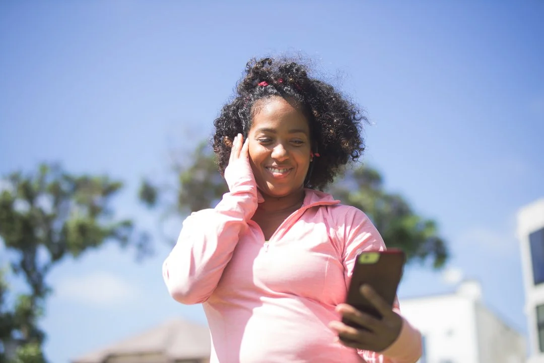 Smiling woman in pink hoodie looking at her phone outdoors with blue sky and trees in background.