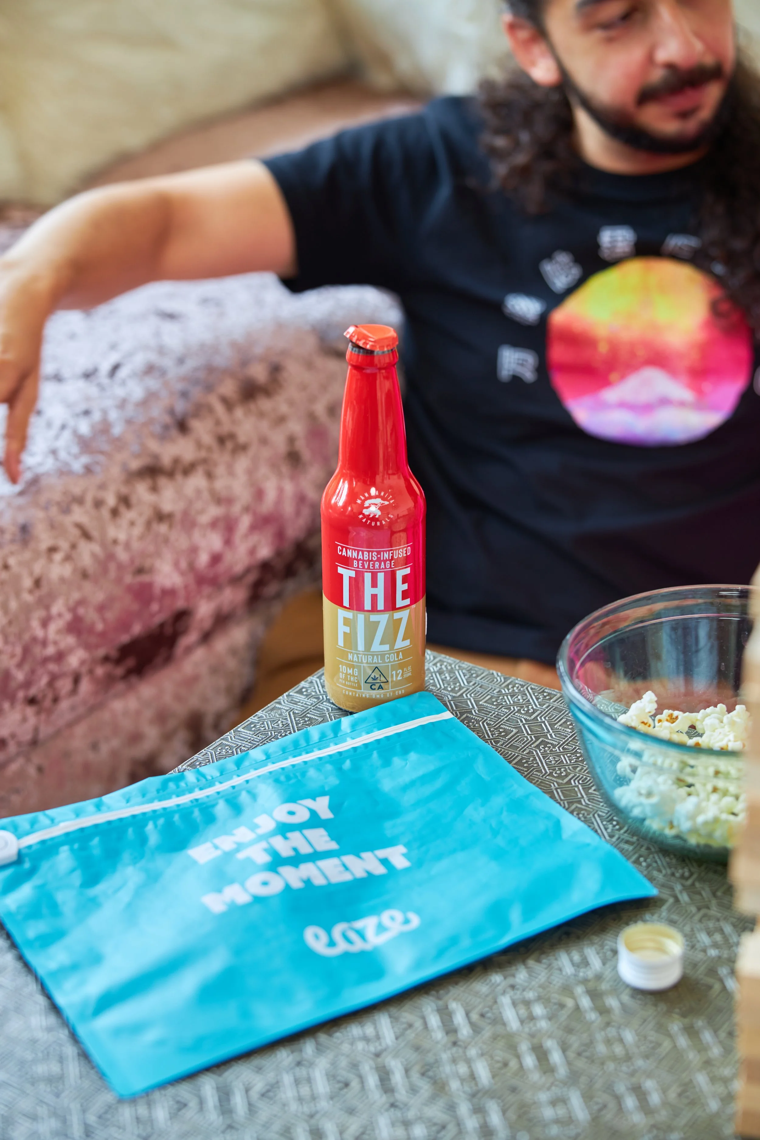 A man sitting at a table with a blue cloth bag that says 'Enjoy the Moment,' a bottle of cannabis-infused natural cola called 'The Fizz,' a bowl of popcorn, and part of a person's arm and face visible in the background.
