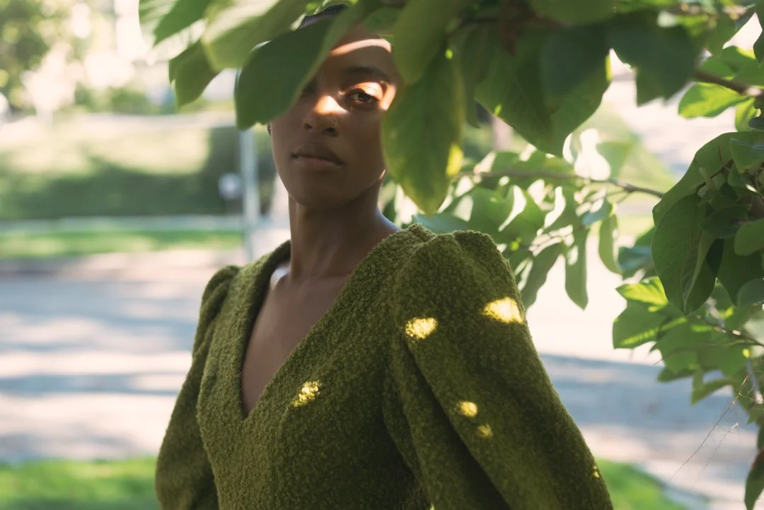 A woman wearing a green textured top standing outdoors partially covered by tree leaves, with sunlight filtering through.