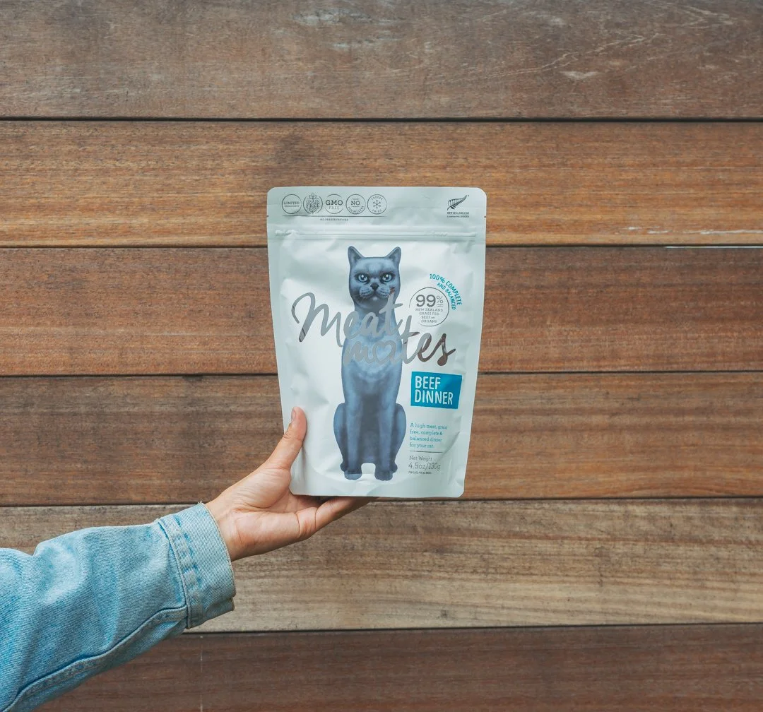 A hand holding a bag of Meow Notes Beef Dinner cat food against a wooden background.
