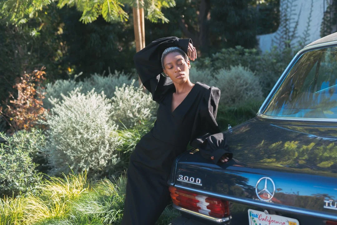 A woman in a black dress standing next to a black Mercedes-Benz 300D car in a garden with lush green plants and trees.
