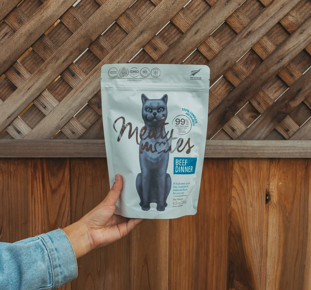 A person holding a bag of meat for cats labeled 'Meaty Mice' with a picture of a black cat, against a wooden fence background.