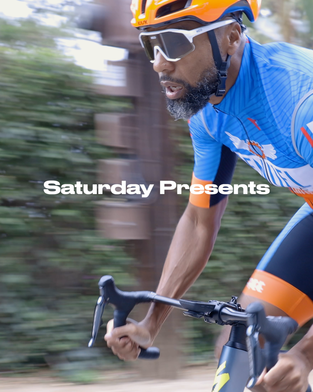 A man cycling outdoors wearing a blue cycling jersey, orange helmet, and sunglasses, with a background of trees and motion blur.