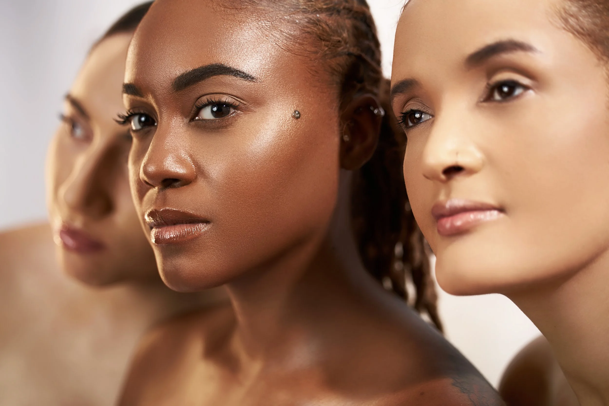 Close-up of three diverse women with natural makeup and confident expressions, standing in a row with neutral backgrounds.