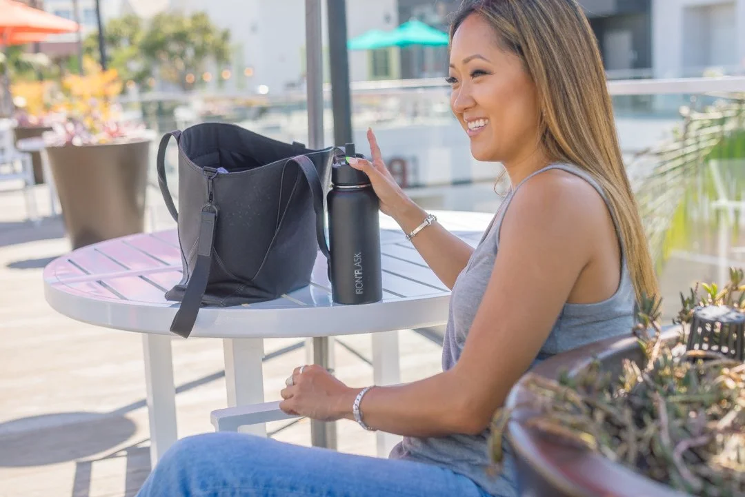 A woman sitting outdoors at a round table, smiling and wearing a gray tank top, with a black bag, a black water bottle labeled 'RONFLASK', and her hand raised to touch a device inside the bag.