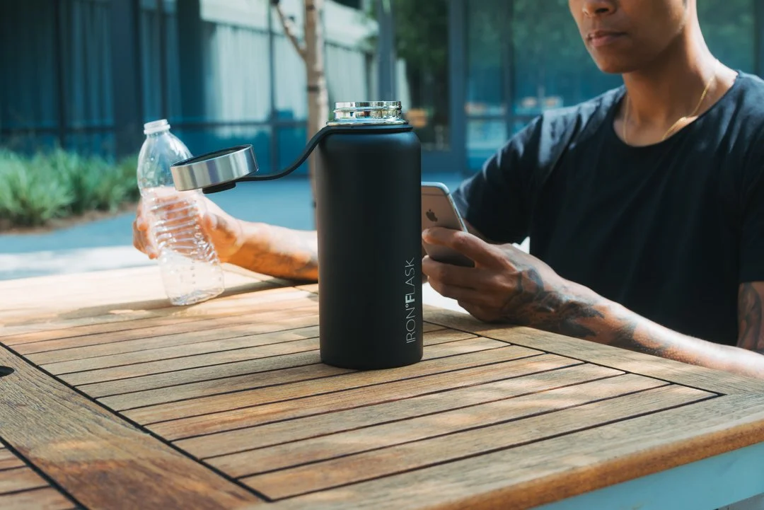 Person sitting at a wooden table outdoors, holding a water bottle and a smartphone, with a black insulated water bottle labeled 'IRONFLASK' on the table.