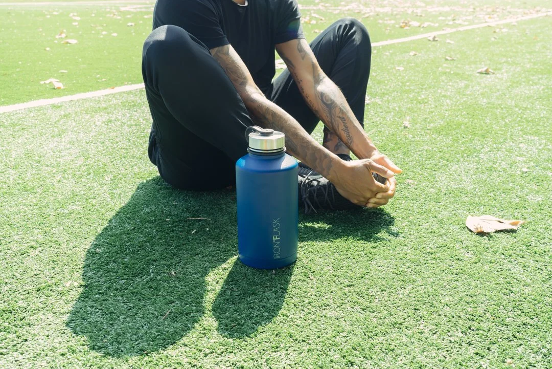 A person sitting on the grass with a blue water bottle nearby, wearing black clothing, on a sunny day.