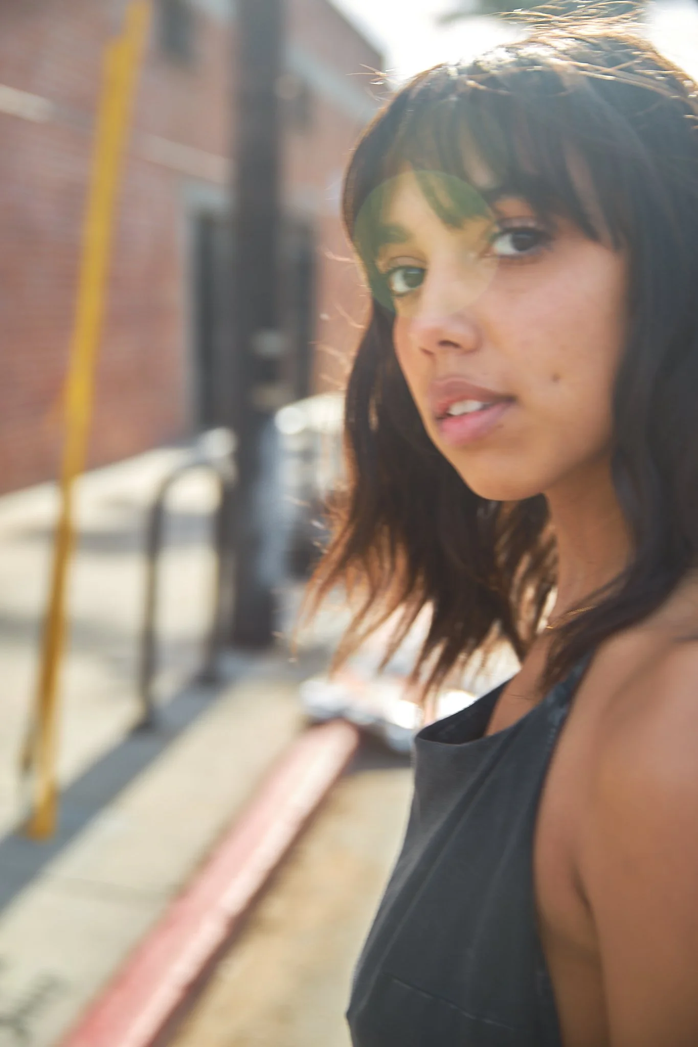 A young woman with dark, shoulder-length hair standing outdoors on a sunny day, with a brick building and a sidewalk in the background.