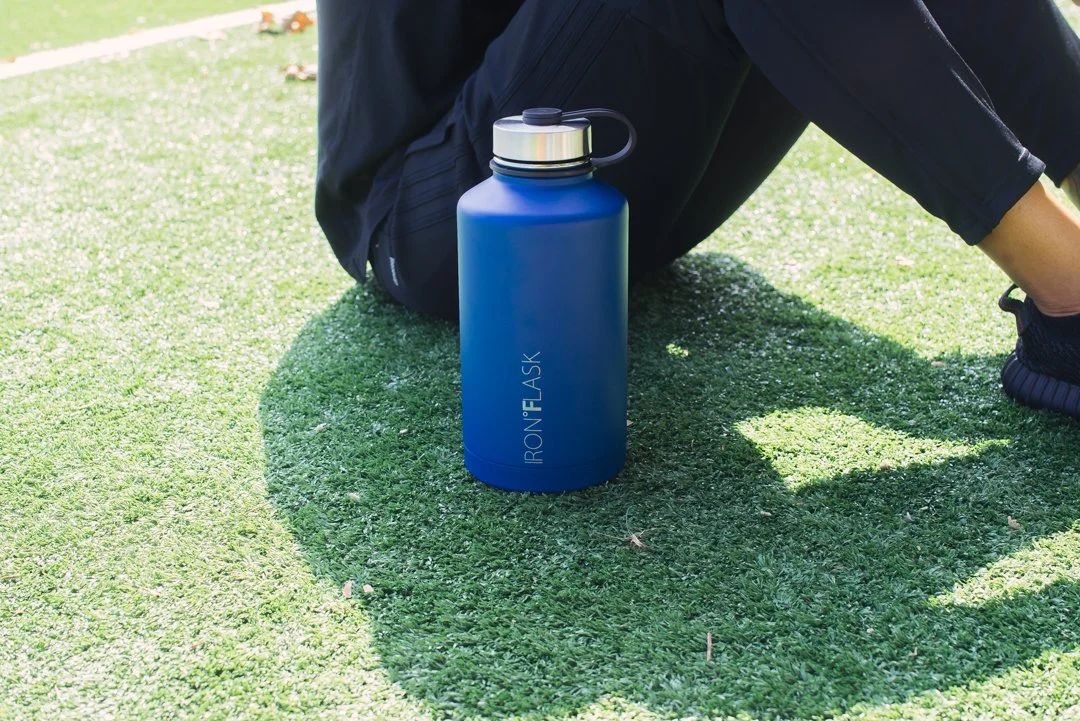 Blue reusable water bottle with silver lid and black handle, labeled 'IRONFLASK', on green grass near seated person wearing black pants and black shoes.