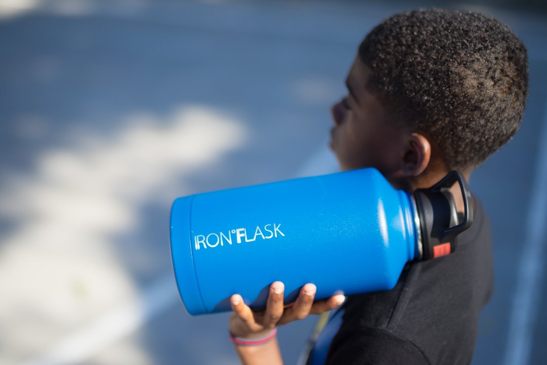 A young boy holding a large blue IRON FLASK water bottle on his shoulder, looking upwards, with a blurred background.