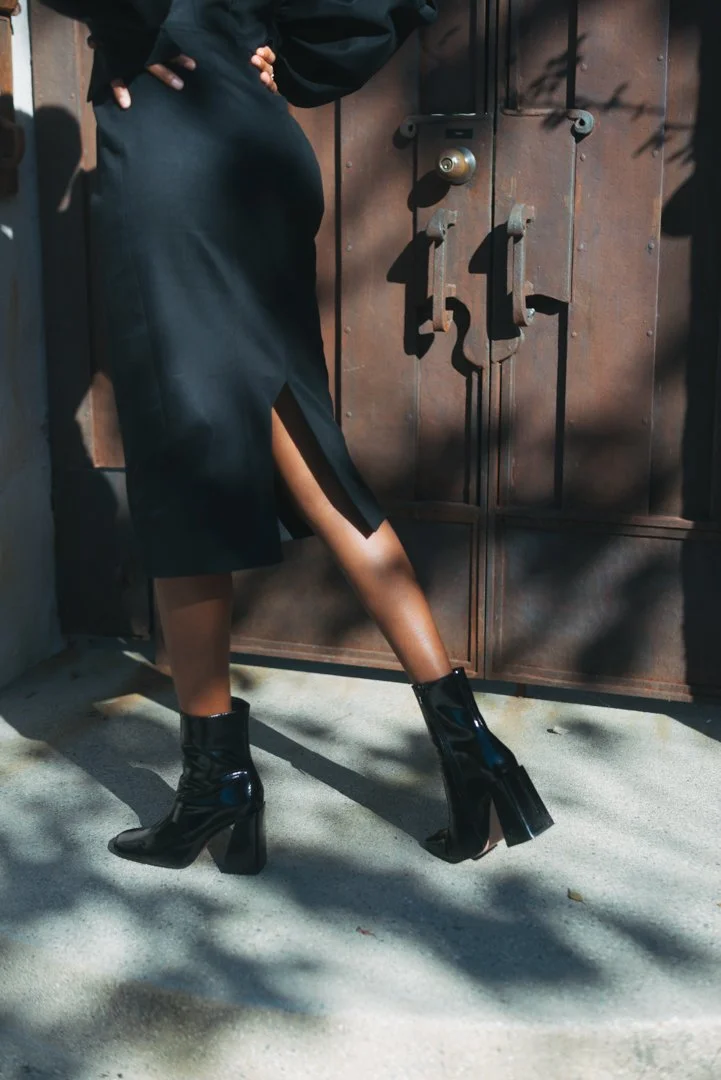 A woman wearing a black dress and shiny black high-heeled boots standing in front of a wooden door with shadows cast on the ground.