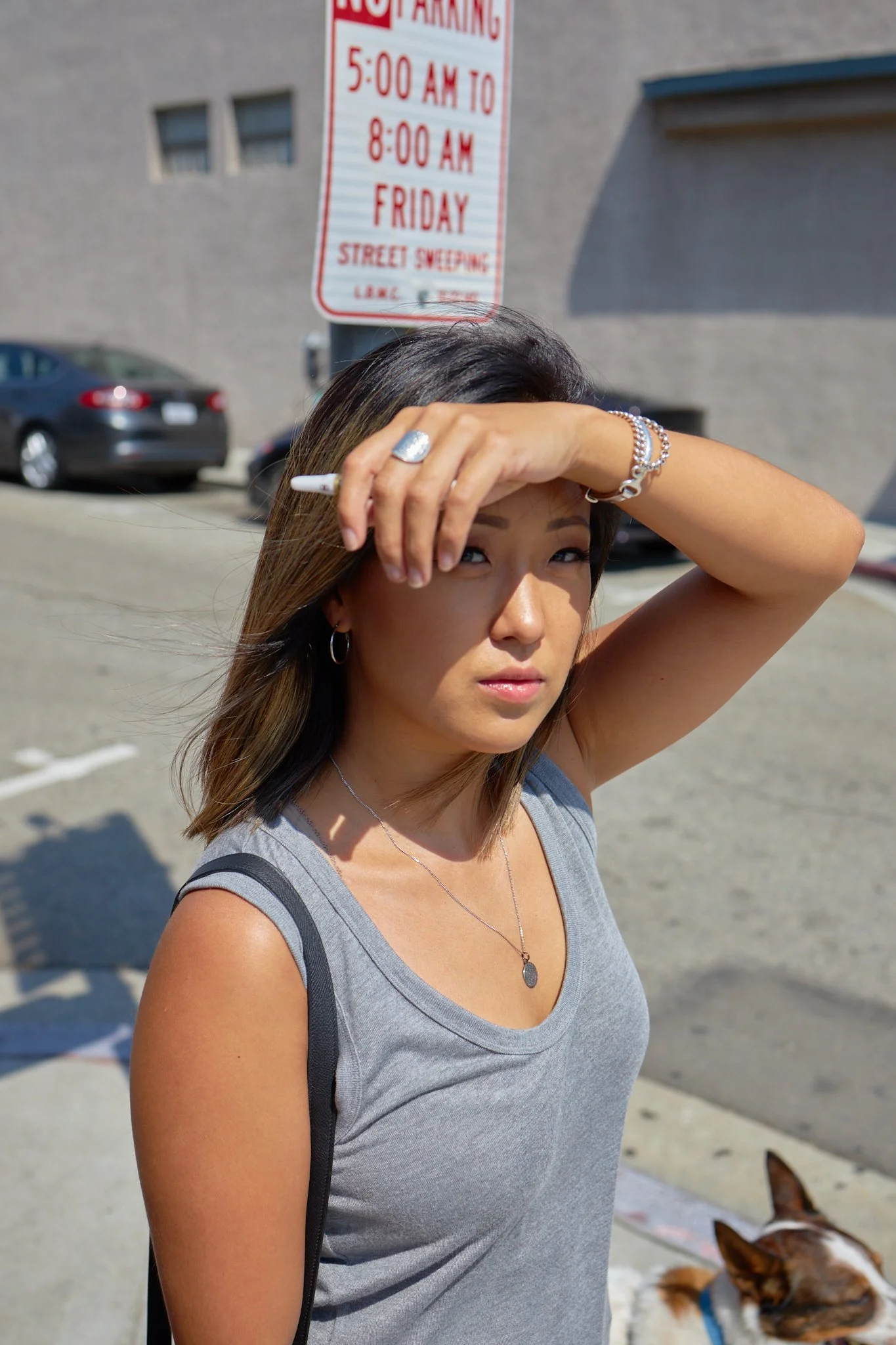 Young woman with brown hair, wearing a gray tank top, jewelry, and holding a cigarette, standing outdoors on a sunny day with a parking lot, cars, and a street sign in the background.
