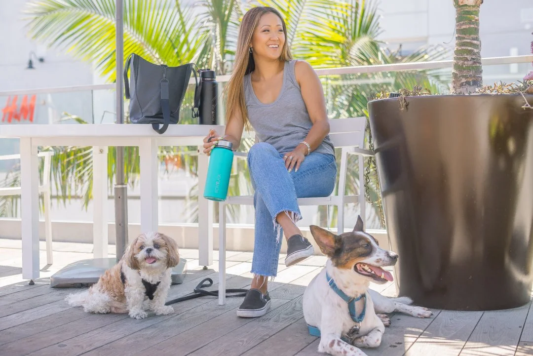 Woman sitting on a chair outdoors, smiling, with two dogs nearby, on a balcony with palm trees in the background.