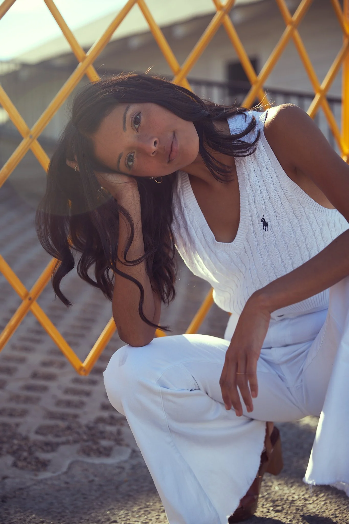 A woman with long dark wavy hair, wearing a white sleeveless knit top and white pants, kneeling on a sandy surface in front of an orange lattice structure, gazing at the camera.
