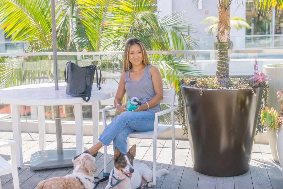 A woman sitting on a patio with two dogs, holding a reusable water bottle, surrounded by potted plants and tropical greenery.