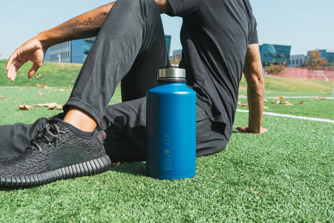 Person sitting on the grass on a sports field, wearing black athletic clothes and sneakers, with a blue water bottle nearby.