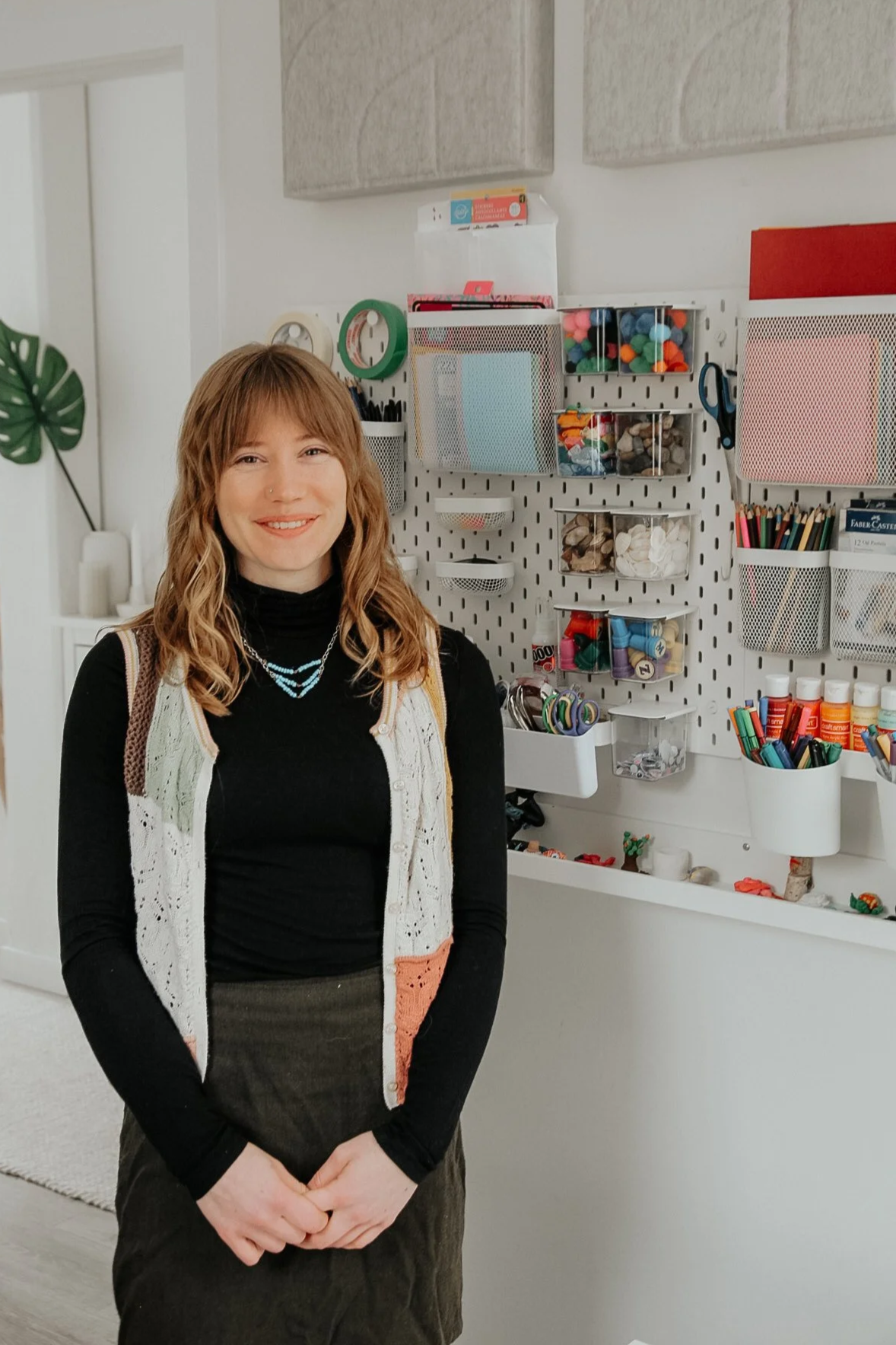A woman with wavy, shoulder-length brown hair, wearing a black turtleneck, a beige cardigan with colored accents, and a dark skirt, standing in front of a craft organizer wall in a room with a large green leaf decoration on the wall.