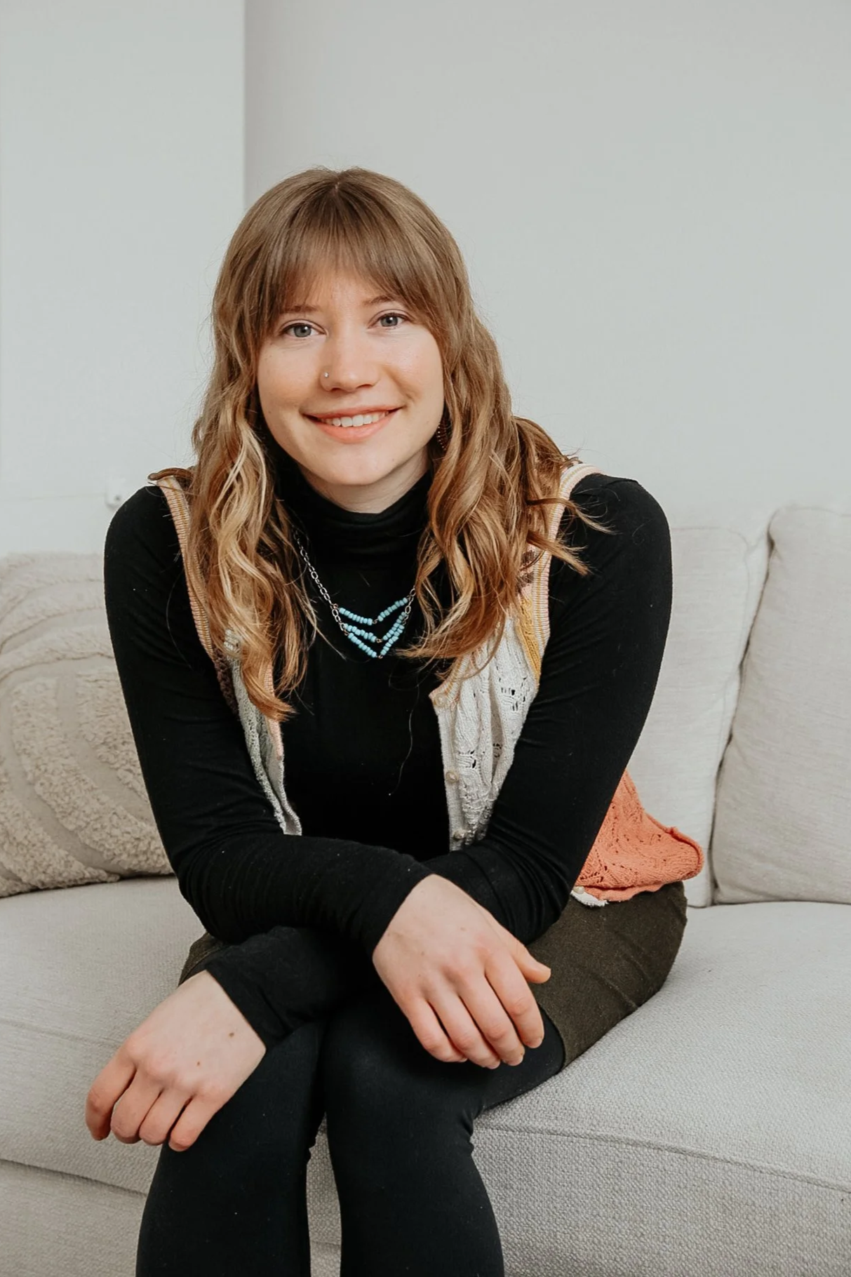 Young adult woman sitting on a beige couch, leaning forward with a kind, warm, and friendly smile on her face.