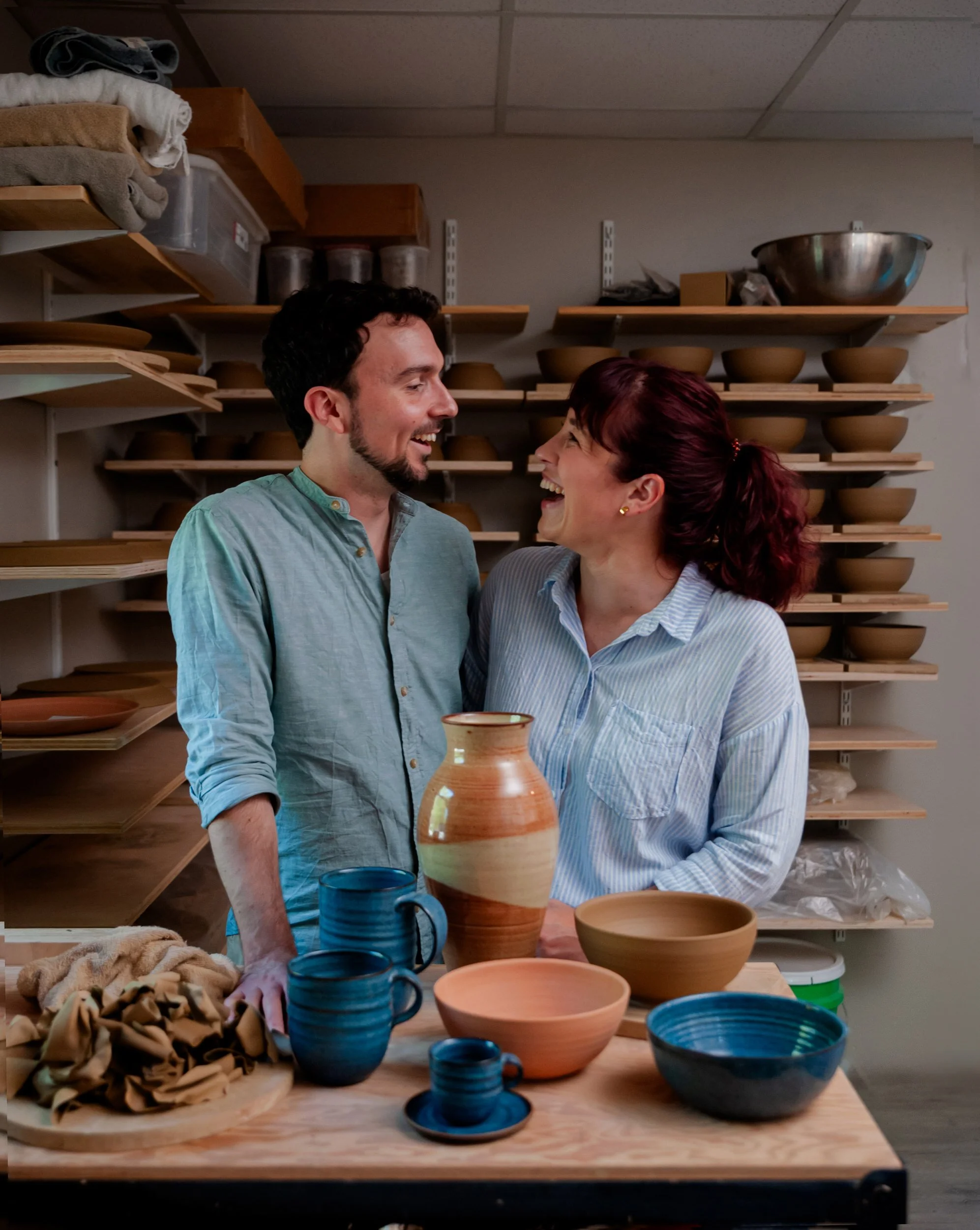 A couple with their arms around each other smiling at each other as they stand in a pottery studio