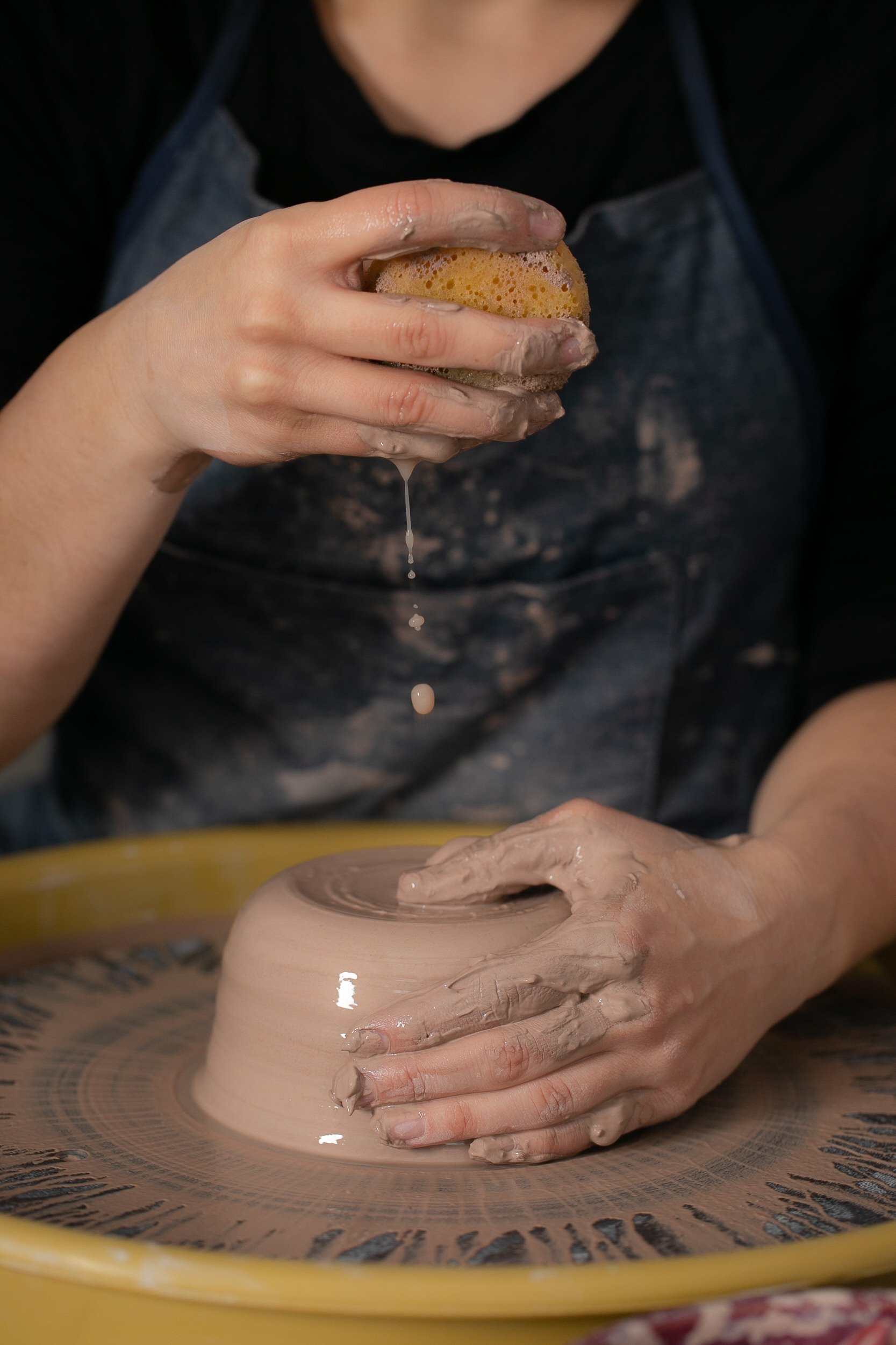 A person squeezing water from a sponge onto a ball of clay on a pottery wheel