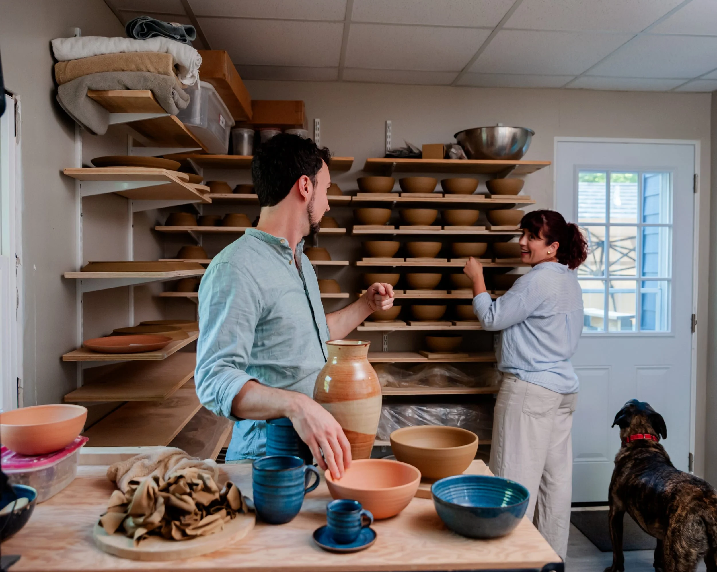 Two people in a pottery studio talking with a dog standing next to them