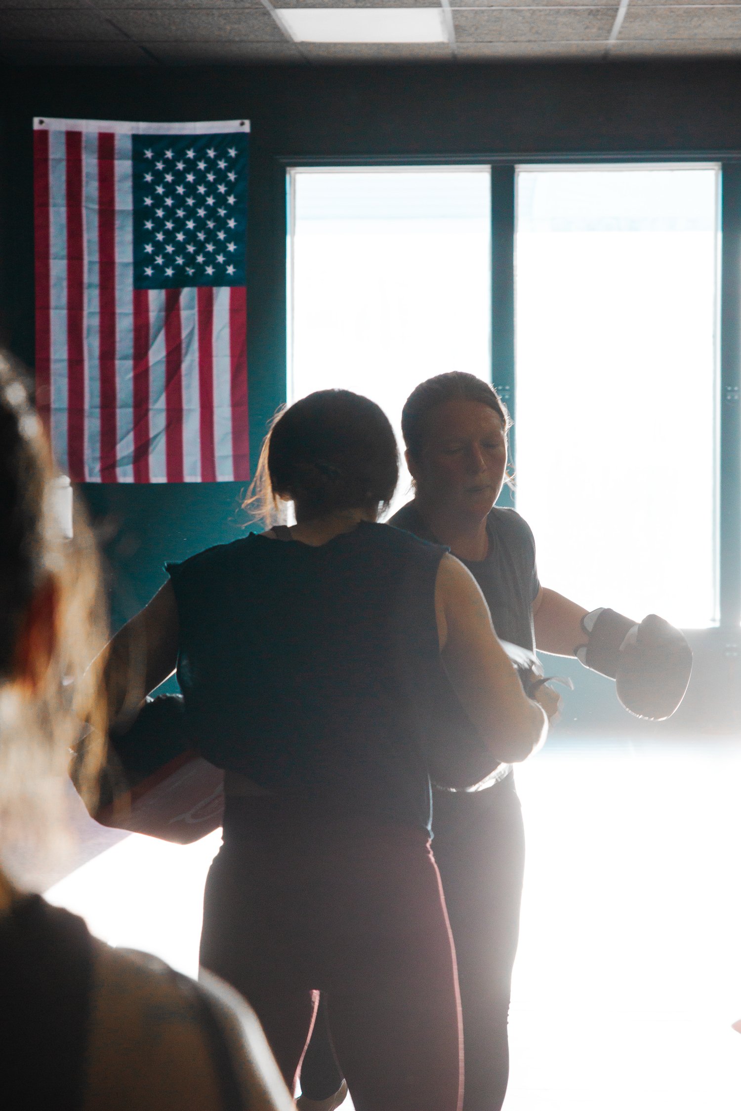 Two women boxing in a gym with an American flag on the wall.