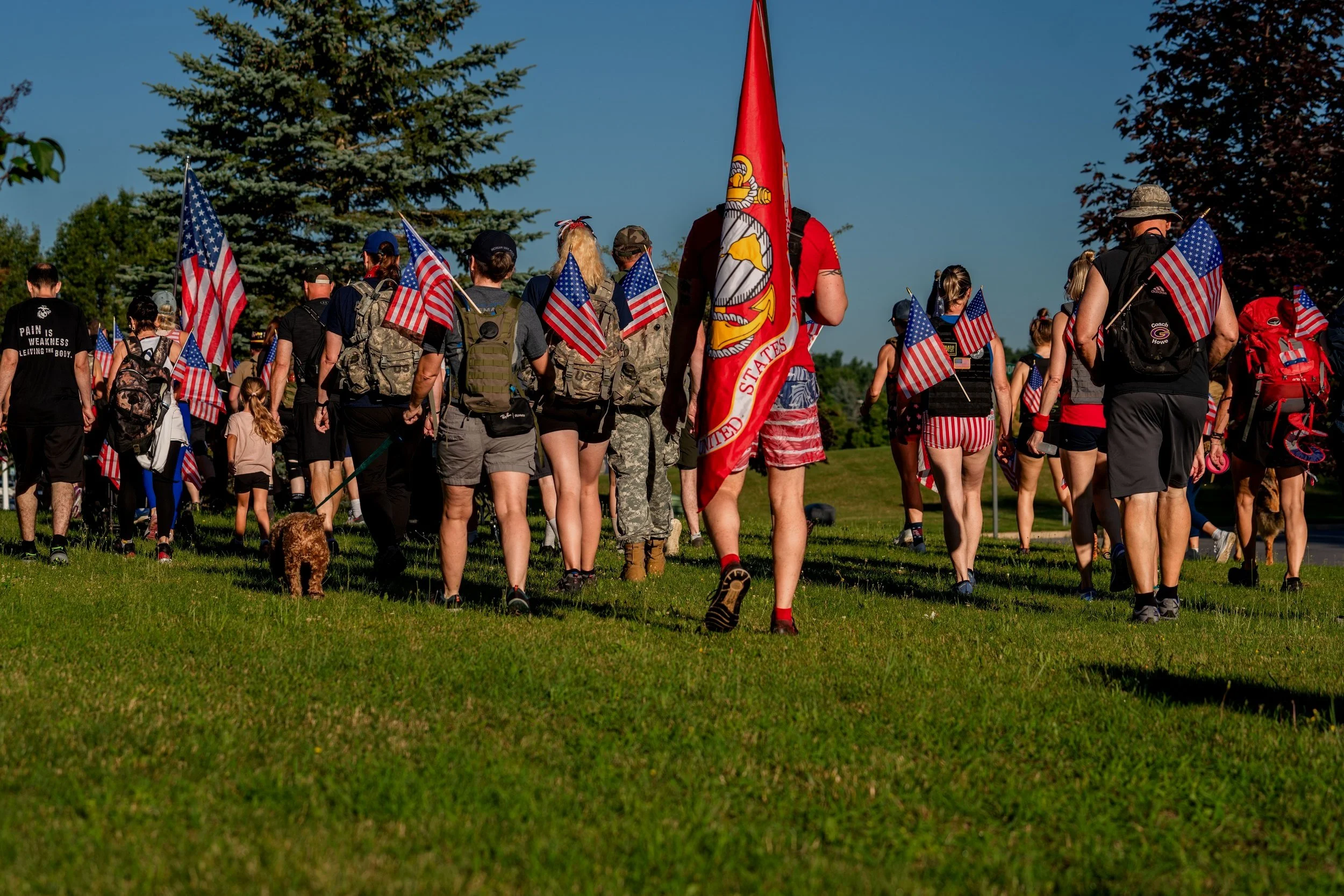 Group of people, some in military attire, carrying American flags and marching in a grassy area, accompanied by a dog.