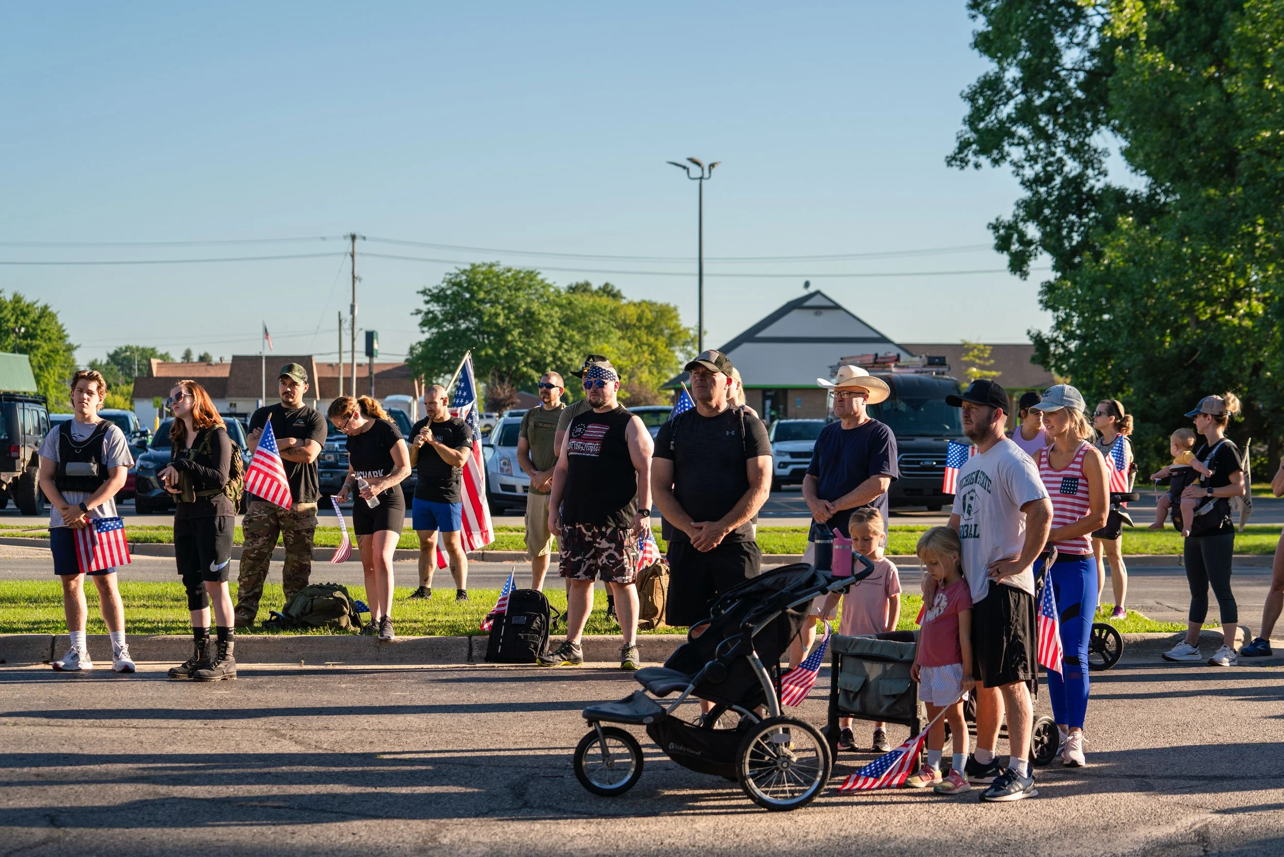 Group of people holding American flags at an outdoor gathering, some with strollers, on a sunny day.