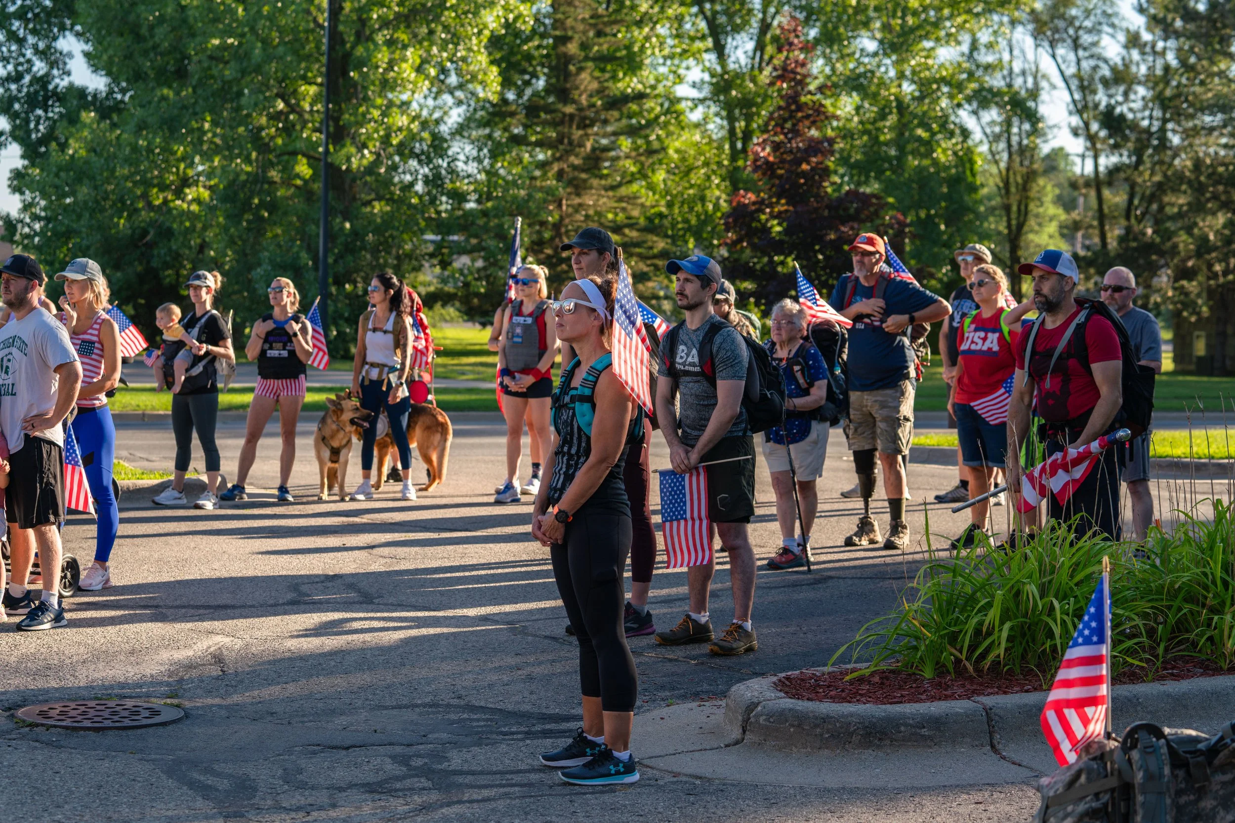 A group of people gathered outdoors holding American flags, some with backpacks and athletic clothing, in a park setting with greenery. It appears to be a patriotic event or gathering.