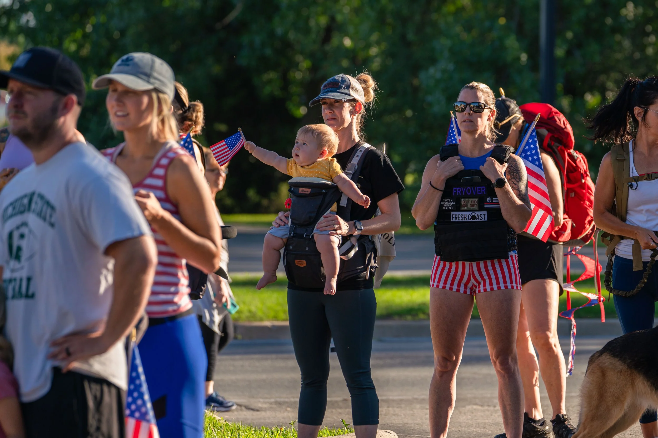 Group of people participating in a parade or event, some holding American flags; includes women wearing shorts and tank tops, a baby in a carrier, and a German Shepherd.