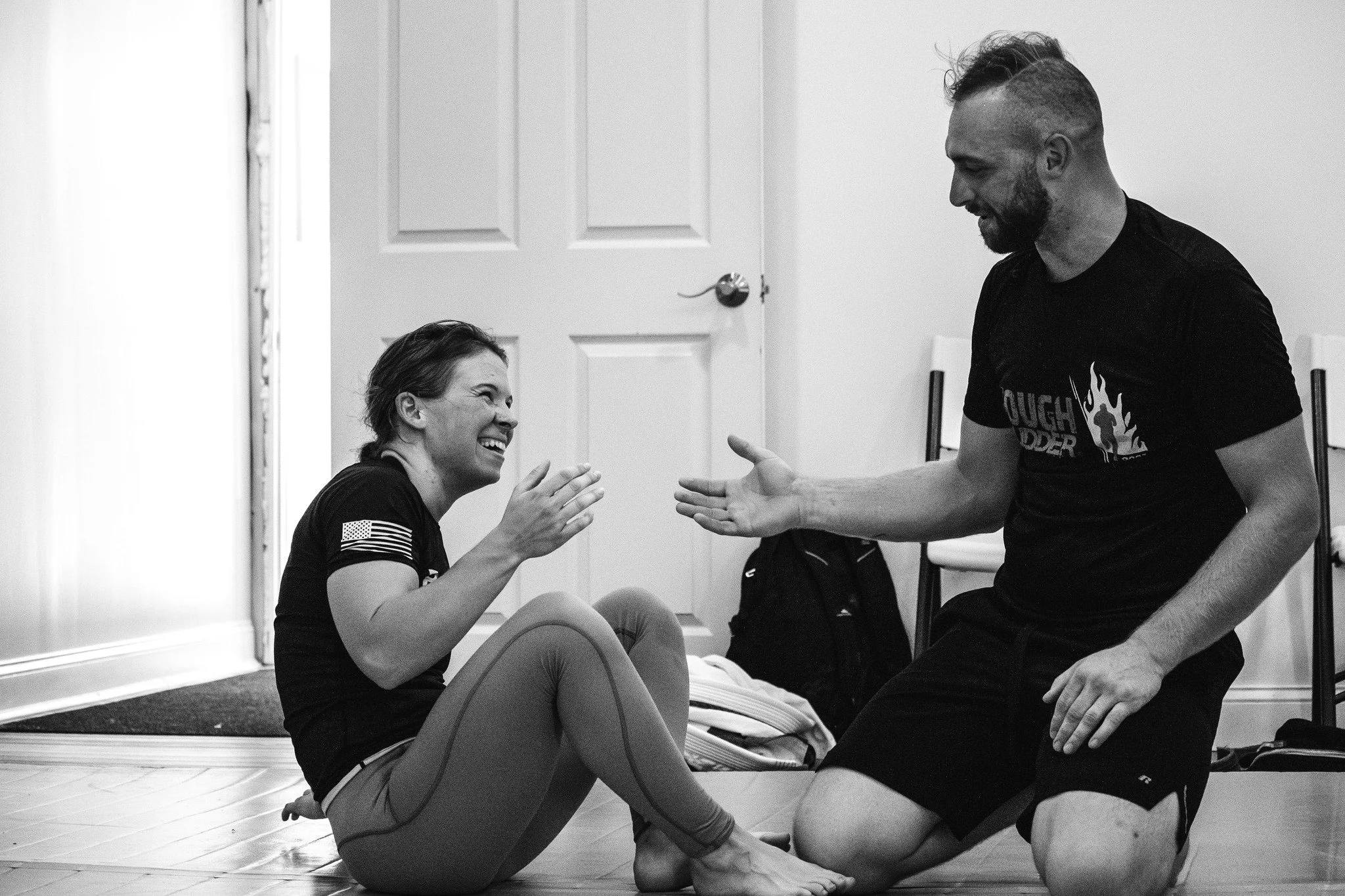 Two people sitting on the floor smiling and reaching out to each other, wearing athletic clothing, in a room with chairs and a door in the background.