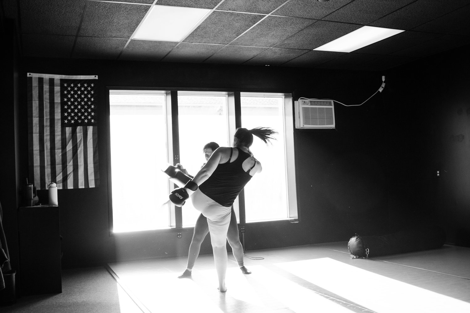 Two women practicing martial arts in a dimly lit gym with an American flag hanging on the wall.