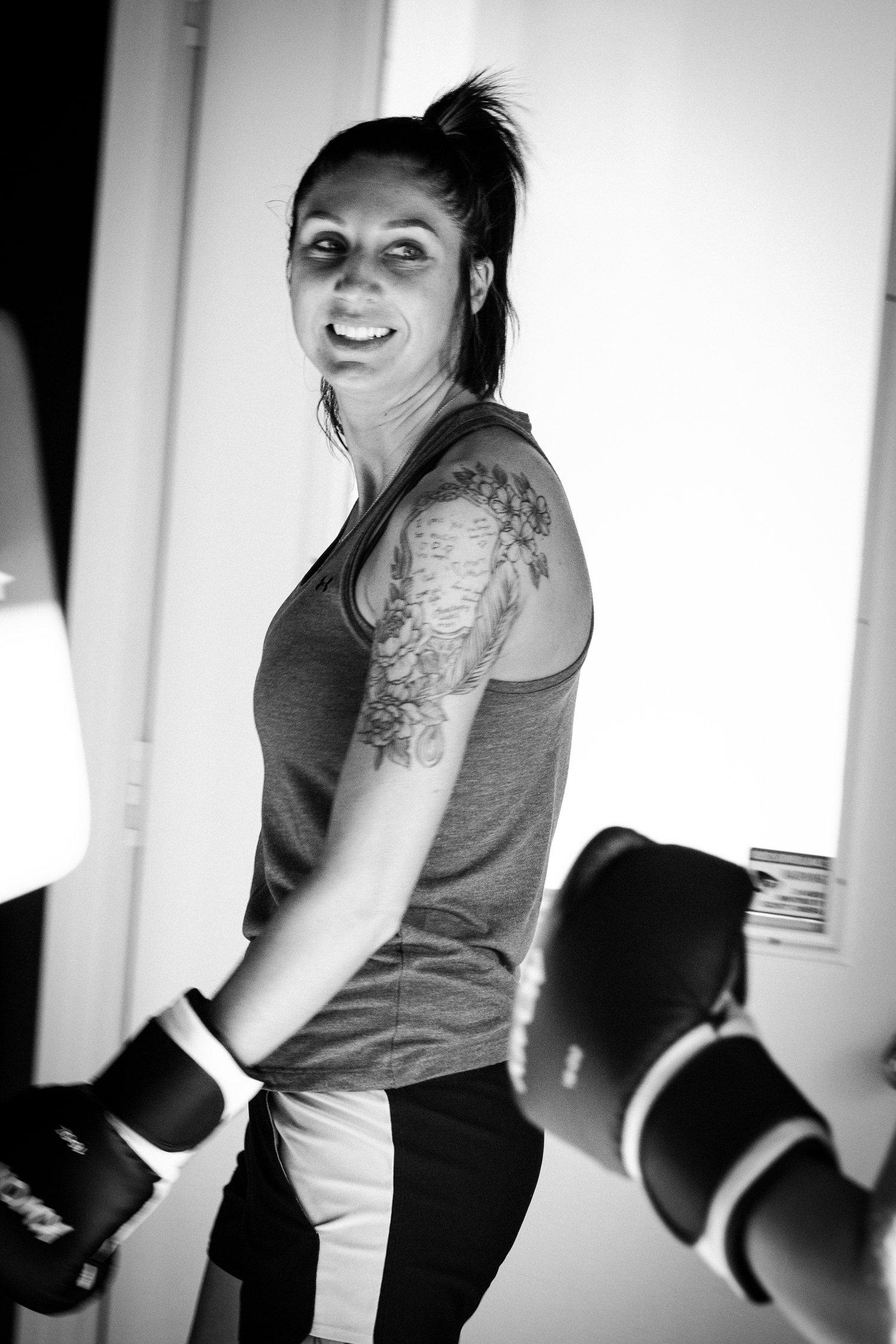 Black and white photo of a woman in a gym wearing boxing gloves and a tank top, with a tattoo on her arm, smiling and looking sideways.