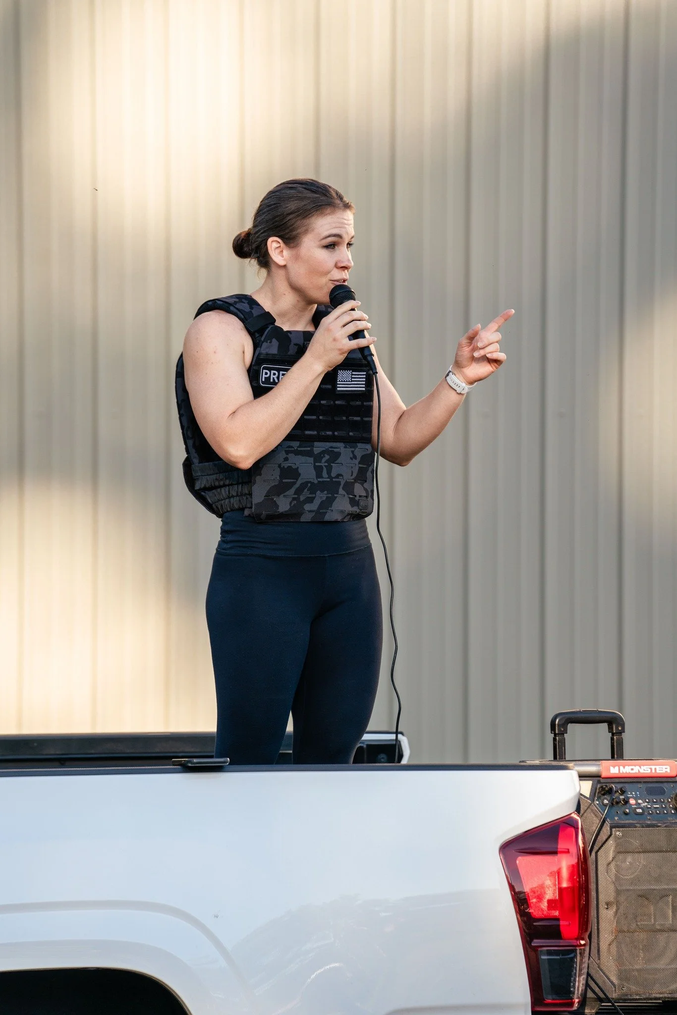 Woman standing in the back of a pickup truck, wearing a tactical vest labeled "PRESS," speaking into a microphone.