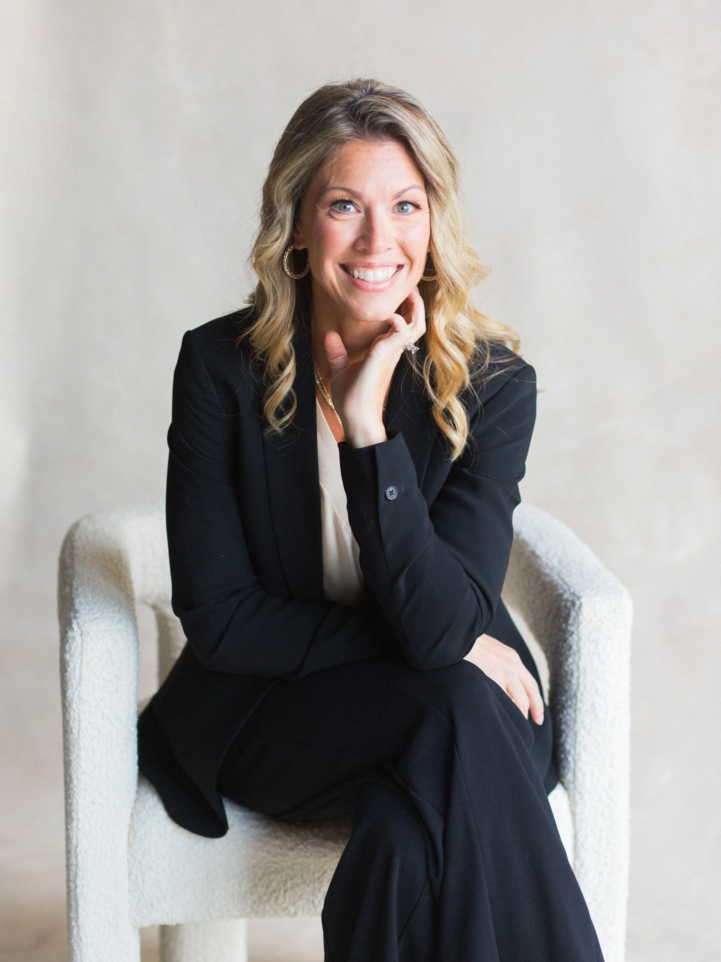 A woman with blonde hair, blue eyes, and a big smile, sitting on a cream-colored textured chair, wearing a black blazer and matching black pants, with hoop earrings and a gold necklace, posing in front of a plain beige background.