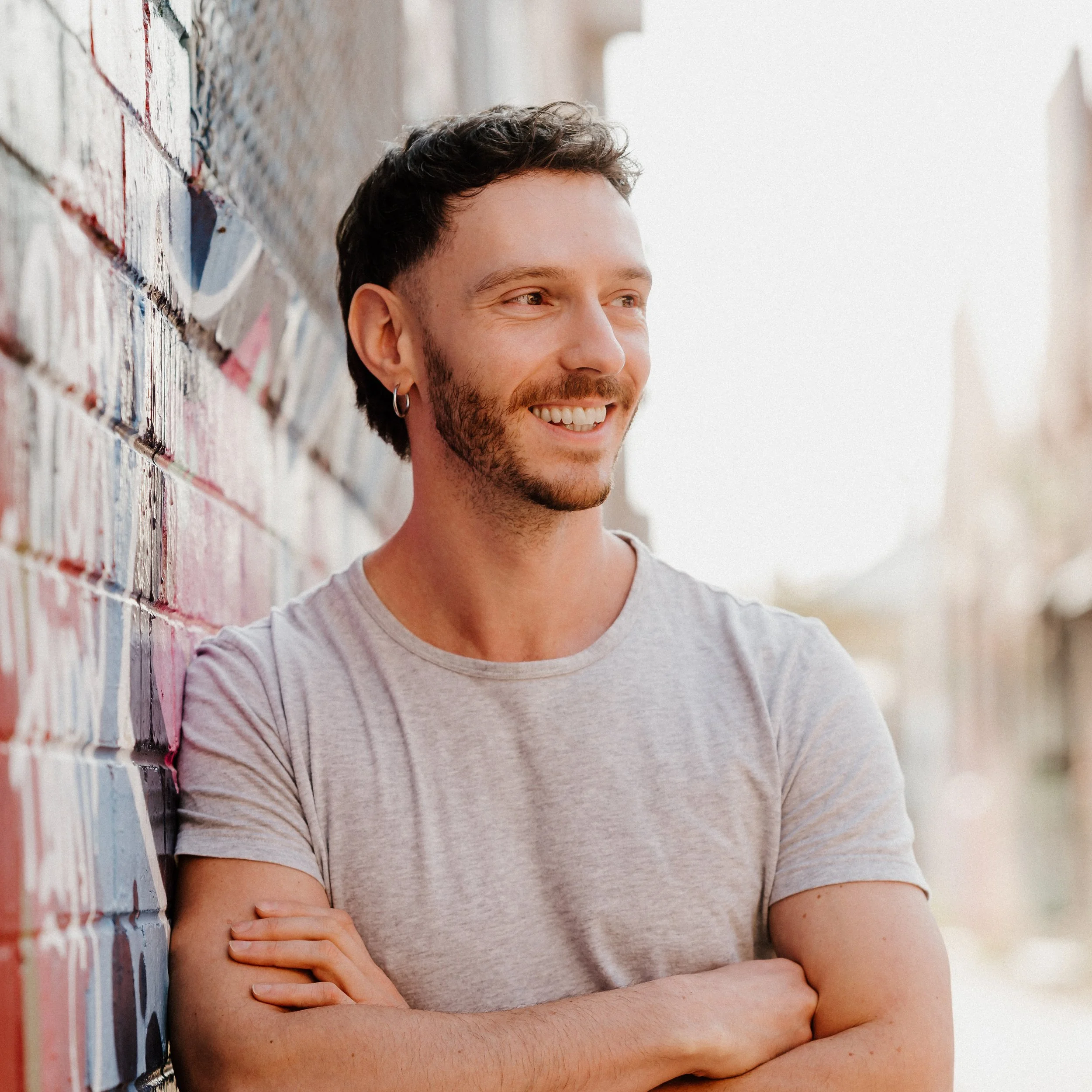 A smiling man with dark hair and a beard, wearing a light gray t-shirt, standing outdoors with arms crossed, leaning against a colorful graffiti-covered brick wall.