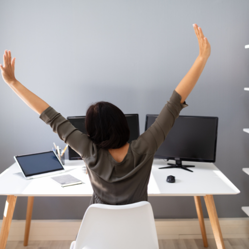 Person with short dark hair sitting at a desk with two computer monitors, raising their arms in the air.