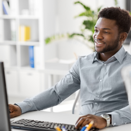 A man sitting at a desk in front of a computer, smiling, in a modern office setting with white furniture and plants in the background.