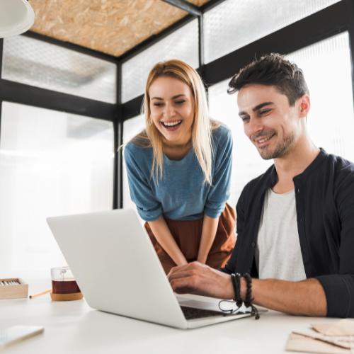 Two young adults, a woman and a man, happily looking at a laptop screen in a modern office setting.