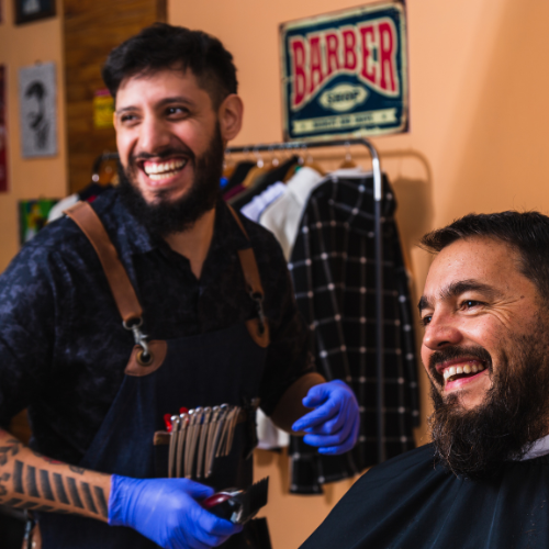 Two smiling men at a barbershop, one standing with an apron and gloves holding scissors, the other sitting in a barber chair. Barber sign and clothing in the background.